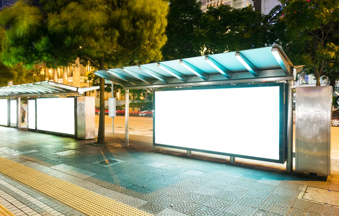 Parada de autobús iluminada con grandes espacios publicitarios en una avenida nocturna.